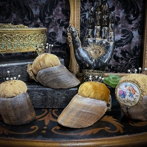 May include: Four antique pincushions made of wood and velvet. The pincushions are decorated with gold trim and have a variety of designs, including a hand with an eye, a floral design, and a miniature painting of a couple.