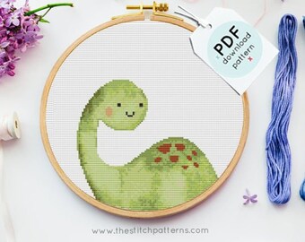 May include: A cross-stitch pattern of a green dinosaur with brown spots, framed in a wooden hoop. The pattern is accompanied by four skeins of embroidery floss in shades of purple, blue, pink, and gray. The text "PDF download pattern" is printed on a white tag.