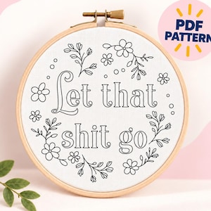 Puede incluir: Un bastidor de bordado circular de madera que contiene tela blanca con un diseño de líneas negras. El diseño presenta motivos florales que rodean el texto Let that shit go. Una insignia circular rosa en la esquina superior derecha muestra el texto PDF PATTERN.