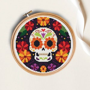 May include: A colorful cross-stitch embroidery of a sugar skull with floral details. The skull is white with black eyes and a red heart on the forehead. The flowers are orange, yellow, red, and purple.