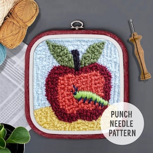May include: Punch needle pattern of a red apple with a green worm on a light blue background. The pattern is framed in a red wooden frame. The text "PUNCH NEEDLE PATTERN" is printed below the frame.
