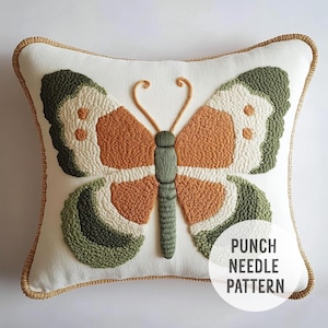 Könnte beinhalten: Ein weißes Kissen mit einem braunen und grünen Schmetterlings-Punch-Needle-Design. Das Kissen hat einen goldenen Rand und der Text "Punch Needle Pattern" ist in der rechten unteren Ecke aufgedruckt.