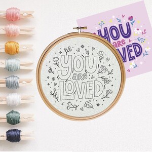 May include: A white embroidery hoop with a black outline of the phrase "You are loved" surrounded by small floral designs.
