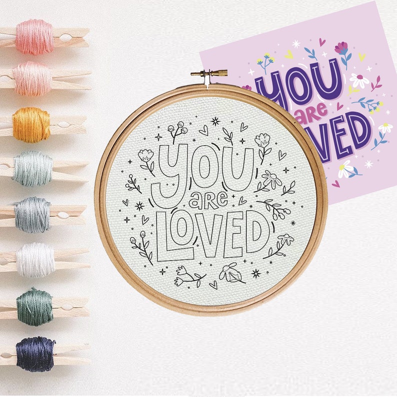 May include: A white embroidery hoop with a black outline of the phrase "You are loved" surrounded by small floral designs.