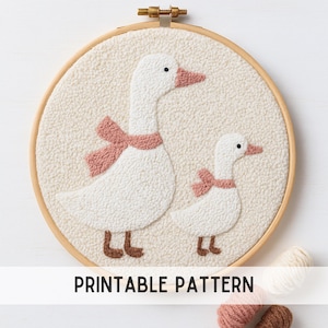 May include: A round, cream-colored textile art piece featuring two white geese with pink bows around their necks. The geese are set against a textured background within a wooden embroidery hoop. The text "PRINTABLE PATTERN" is at the bottom.