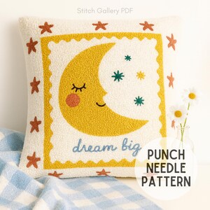 May include: Cream-colored square pillow with a yellow crescent moon face, surrounded by orange stars. The phrase "dream big" is embroidered in blue. The pillow is a punch needle pattern.