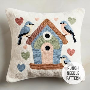 May include: A white pillow with a punch needle design of a blue birdhouse with two birds perched on the sides and hearts scattered around. The text "PUNCH NEEDLE PATTERN" is in the bottom right corner.
