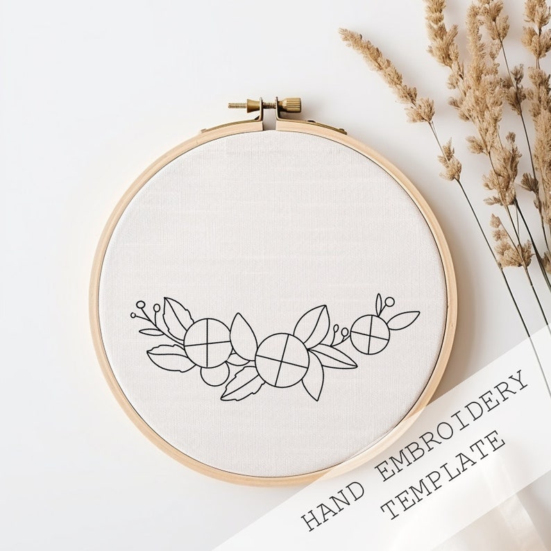 May include: A wooden embroidery hoop with cream fabric and a black floral design. The text "HAND EMBROIDERY TEMPLATE" is printed on a white paper. Dried plants are in the background. The hoop is a popular craft item.