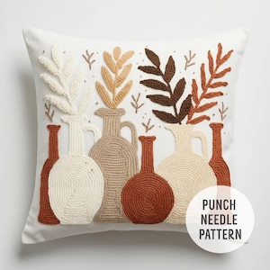 May include: A white pillow with a punch needle embroidery design of brown, beige, and cream colored vases with leaves. The text "PUNCH NEEDLE PATTERN" is in the bottom right corner of the pillow.