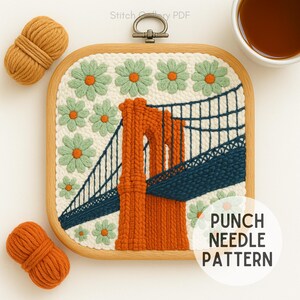 May include: A square, wooden embroidery hoop with a punch needle pattern of the Brooklyn Bridge in orange and navy blue, surrounded by green and orange flowers. The text "PUNCH NEEDLE PATTERN" is in a white circle. Two bundles of orange yarn are on the left.
