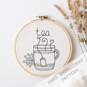 Peut inclure: Motif de broderie noir et blanc d'une tasse de thé avec de la vapeur et un sachet de thé, avec le mot "tea" au-dessus. Le motif est sur un tissu blanc dans un cerceau de broderie en bois.