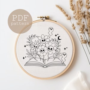 May include: Black and white embroidery pattern of a book with flowers and skulls. The flowers have skulls in the center and are surrounded by leaves. There are two ghosts on either side of the book. The pattern is on a white fabric in a wooden embroidery hoop.