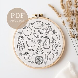 May include: A black and white embroidery pattern of various fruits and flowers in a circle on a white fabric background. The pattern includes a pineapple, apple, pear, watermelon, lemon, orange, cherries, kiwi, plums, and bananas.
