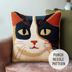 May include: A square cushion with a cat face design in orange, navy blue, yellow, and red. The cushion has a white background and the text "PUNCH NEEDLE PATTERN" in a white circle. The cushion is a decorative item.