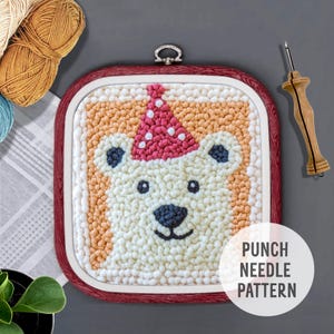 May include: A punch needle pattern featuring a white polar bear wearing a red and white party hat. The pattern is framed in a red wooden hoop. The text "PUNCH NEEDLE PATTERN" is printed below the hoop.