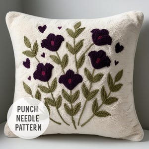 May include: A white pillow with a punch needle embroidery design of purple flowers and green leaves. The pillow has the text "Punch Needle Pattern" on the bottom left corner.