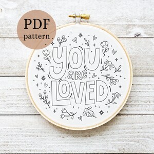 May include: A white embroidery hoop with a black outline of the phrase "you are loved" surrounded by small floral designs. The hoop is on a white wooden background.
