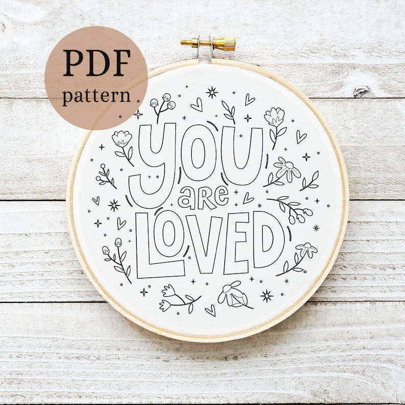 May include: A white embroidery hoop with a black outline of the phrase "you are loved" surrounded by small floral designs. The hoop is on a white wooden background.