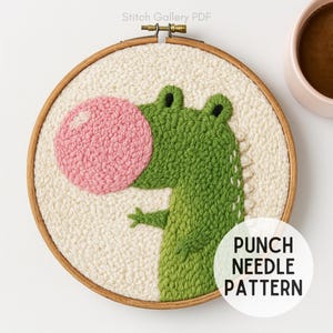 Op de afbeelding: Een punch needle patroon met een groene alligator die een roze bel blaast. De alligator staat tegen een crèmekleurige achtergrond in een houten borduurring. De tekst "PUNCH NEEDLE PATTERN" staat in een witte cirkel.