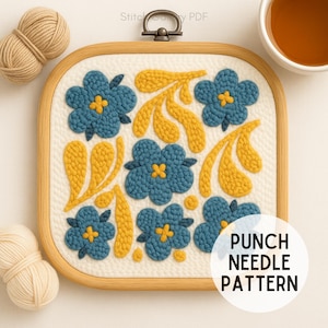 Op de afbeelding: Een vierkante houten borduurring met een bloemenpatroon. Het ontwerp toont blauwe bloemen met gele centra en gele bladeren. De tekst "PUNCH NEEDLE PATTERN" staat in een witte cirkel. Garen en een kopje thee zijn ook zichtbaar.