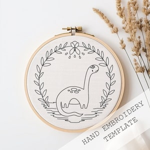 May include: A black and white line drawing embroidery template of a dinosaur with a long neck standing in a wreath of leaves. The template is on a white fabric background in a wooden embroidery hoop. The text "HAND EMBROIDERY TEMPLATE" is written in black on a white background below the hoop.