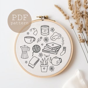 May include: A black and white embroidery pattern featuring a variety of cozy and relaxing items, including a cup of tea, a book stack, a candle, a potted plant, a teapot, a sleep mask, yarn, and a pair of socks. The pattern is displayed on a white fabric background inside a wooden embroidery hoop.
