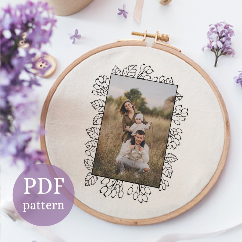May include: A black and white embroidery pattern of a family of three in a field, surrounded by a floral border. The pattern is on a white fabric in a wooden embroidery hoop.