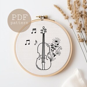 以下が含まれることがあります： バイオリン、音符、花が描かれた白黒の刺繍パターン。パターンは丸い刺繍枠に入っています。