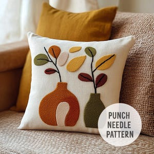 Op de afbeelding: Een wit kussen met een punch needle borduurwerk ontwerp van twee vazen met bladeren in tinten van groen, bruin, oranje en geel. De tekst "PUNCH NEEDLE PATTERN" is zichtbaar in de rechter benedenhoek van de afbeelding.