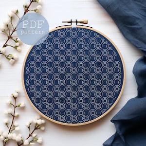 Könnte beinhalten: Ein marineblauer Stickrahmen mit einem weißen Spiralmuster. Der Text "PDF pattern" befindet sich in einem hellblauen Kreis. Der Holzrahmen liegt auf einer weißen Oberfläche mit weißen Blumen und einem blauen Stoff.