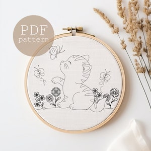 以下が含まれることがあります： 蝶を見上げる猫の白黒線画。猫は花と蝶に囲まれています。絵は木製の刺繍枠に入れた白い布に描かれています。画像の左上隅には茶色の円の中に「PDF pattern」という文字があります。