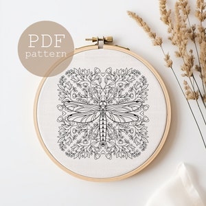 Puede incluir: Patrón de bordado en blanco y negro de una libélula rodeada de diseños florales. El patrón se muestra en un aro de bordado de madera.