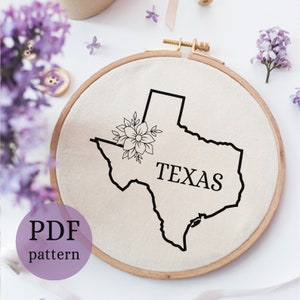 May include: A black and white embroidery pattern of the state of Texas with a floral design and the word "TEXAS" in the center. The pattern is on a white fabric in a wooden embroidery hoop.