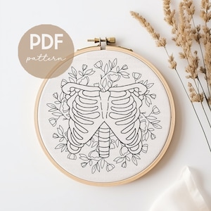 Puede incluir: Un dibujo en blanco y negro de una caja torácica rodeada de diseños florales. El diseño está en una tela blanca en un aro de bordado de madera. El texto "PDF pattern" está en la esquina superior izquierda de la imagen.