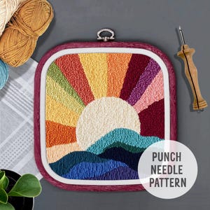 May include: A colorful rainbow sunset punch needle pattern with a white sun and blue waves. The pattern is in a wooden embroidery hoop with a red wood finish. The text "PUNCH NEEDLE PATTERN" is in white text on a black circle.