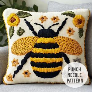 May include: A white cushion with a punch needle embroidery design of a yellow and black bee surrounded by yellow flowers. The text "PUNCH NEEDLE PATTERN" is in the bottom right corner of the cushion.