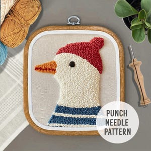 Op de afbeelding: Een ingelijst punch needle patroon van een witte vogel met een rode hoed en een blauw-wit gestreept shirt. Het patroon is gelabeld "Punch Needle Pattern".