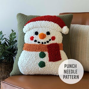 May include: A green pillow with a white snowman design. The snowman has a red hat, orange carrot nose, black eyes, and a red and orange scarf. The text "PUNCH NEEDLE PATTERN" is on the pillow.