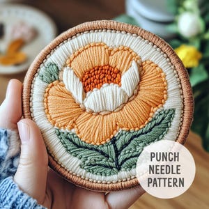 May include: A punch needle pattern featuring a large orange flower with a white center and green leaves. The pattern is circular and has a brown border. The text "Punch Needle Pattern" is printed below the pattern.