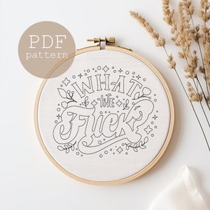 Puede incluir: Un patrón de bordado en blanco y negro para un proyecto de arte de aro. El patrón presenta la frase "What the Fuck" en una fuente estilizada con acentos florales.