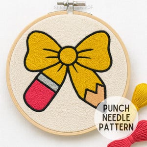 May include: A yellow bow and pencil design is punch needle embroidered on a cream-colored fabric, set in a wooden hoop. The text "PUNCH NEEDLE PATTERN" is displayed in a white circle. The design includes red, yellow, and peach colors.