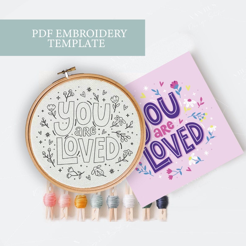May include: A PDF embroidery template featuring the phrase "You are loved" surrounded by flowers in a hoop. The design is also shown on a pink background with purple lettering.