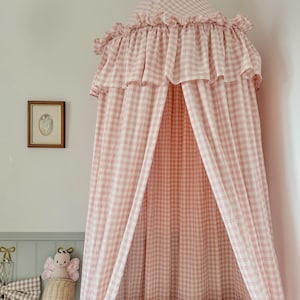 Puede incluir: Un dosel a cuadros rosa y blanco cuelga sobre una cuna de madera. El dosel tiene un borde con volantes y está abierto por delante. Una imagen enmarcada y un peluche en una cesta están en la pared.