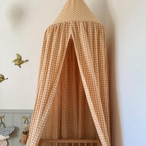 May include: A yellow and white gingham canopy hangs over a wooden crib. The canopy is cone-shaped and drapes down, creating a cozy space. The crib has a patterned pillow inside. Decorative gold birds are mounted on the wall.