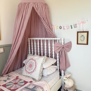 May include: A child's bed with a white frame, featuring a canopy and a large bow made of red and white gingham fabric. The bed is dressed with a floral quilt, decorative pillows, and a white bunny figurine on stacked boxes.