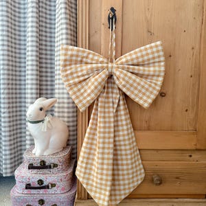 May include: A large, decorative bow made of yellow and white gingham fabric hangs from a black hook on a wooden cabinet. A white ceramic bunny sits atop a stack of floral patterned suitcases. Gingham curtains are in the background.