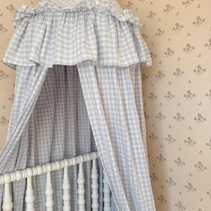 May include: A light blue and white gingham canopy featuring a conical top with a ruffled edge and long, flowing fabric panels that drape downwards. The canopy is positioned above a white wooden cot with a spindle design and a light grey pillow.
