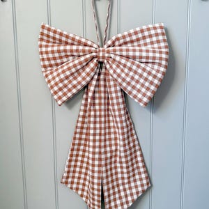May include: A large bow made from brown and white gingham fabric, hanging on a light blue wall. The bow has a long, pointed tail and is suspended by a matching fabric loop. The bow is a decorative item.