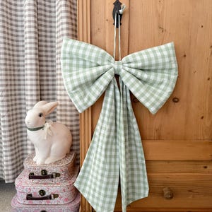 Puede incluir: Un gran lazo de cuadros vichy verde claro y blanco colgado de un armario de madera. Un conejo de cerámica blanca se sienta sobre tres maletas con estampado floral. Una cortina a cuadros grises y blancos está en el fondo.
