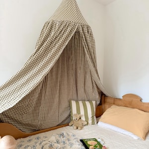 Peut inclure: Une chambre d'enfant avec un lit en bois et un baldaquin à carreaux. Le lit est fait avec une couette matelassée blanche, un oreiller à rayures et un oreiller orange clair. Une voiture jouet en bois et un livre sont également sur le lit.